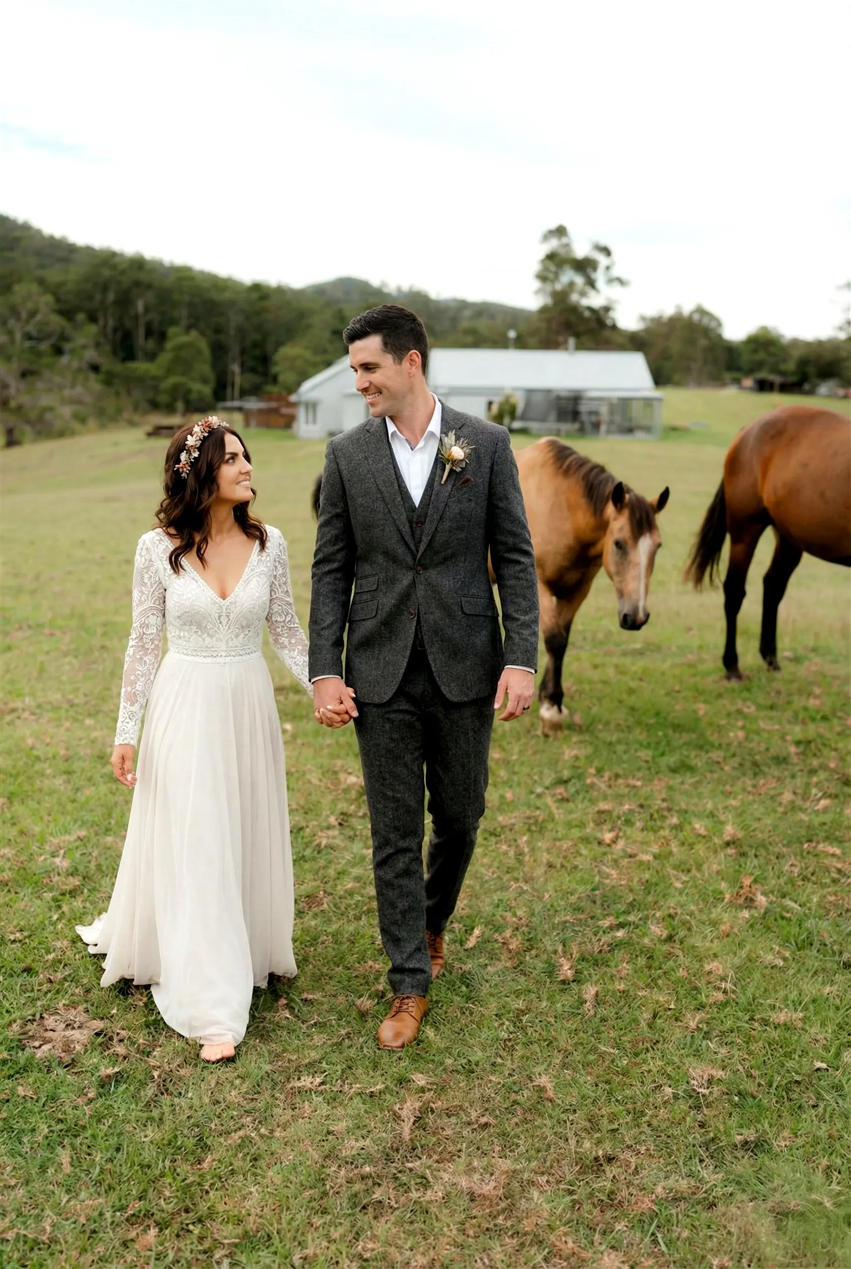 A wedding couple walking hand-in-hand through a vibrant green paddock with horses grazing in the background during a sunset portrait session at Cowbell Creek.