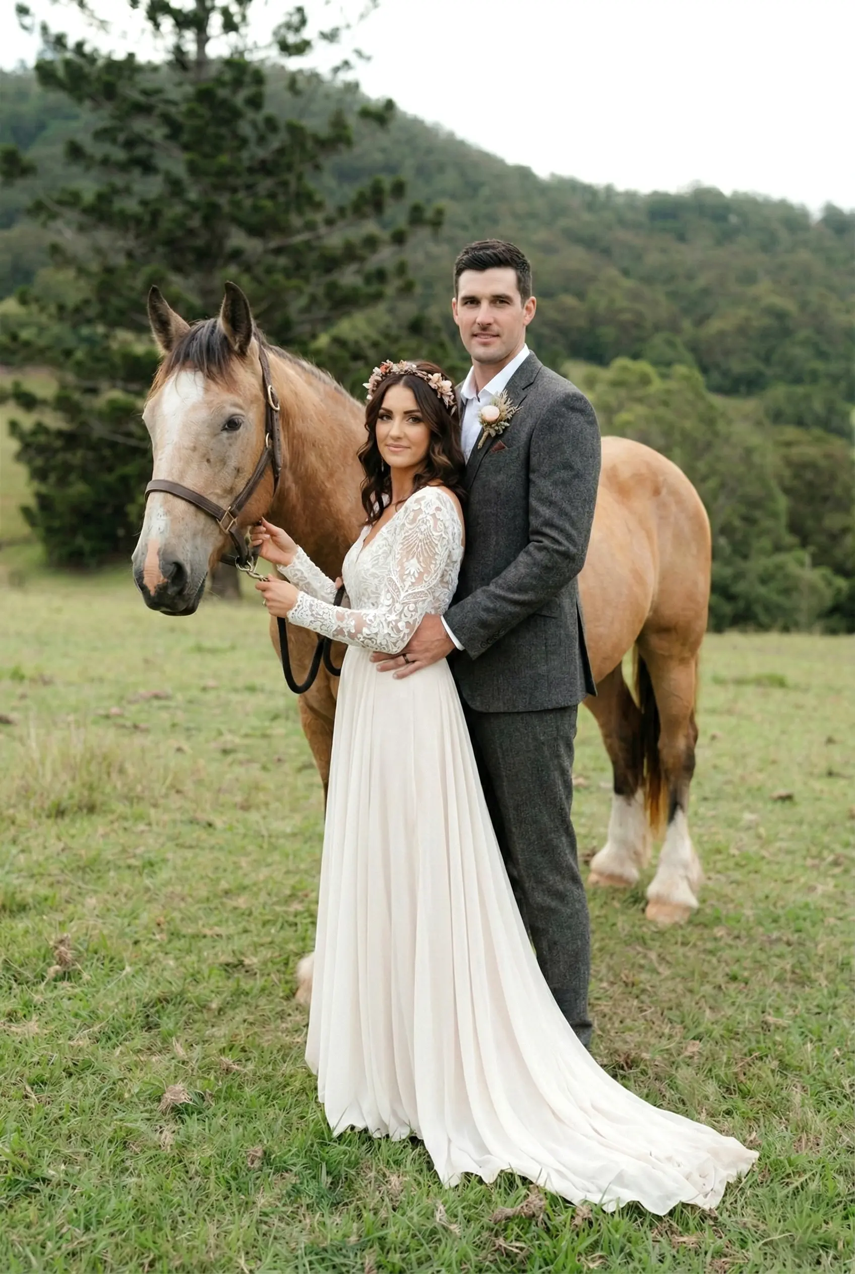 A bride wearing a floral crown and lace gown stands beside a groom in a grey suit, posing with a large tan horse in a rural field at Cowbell Creek wedding venue.