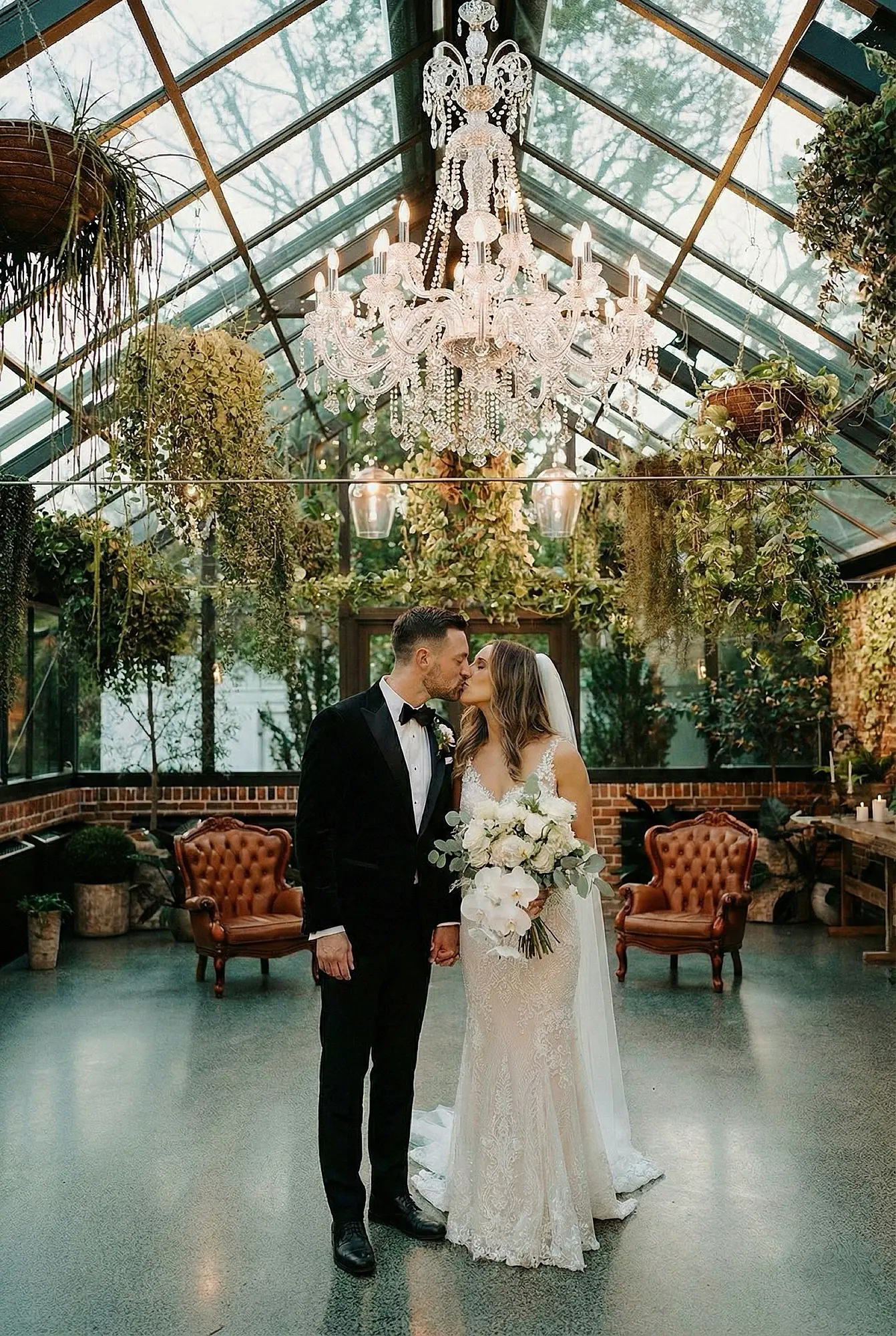 Bride and groom standing under a crystal chandelier in The Glasshouse at The Valley Estate, surrounded by lush greenery.