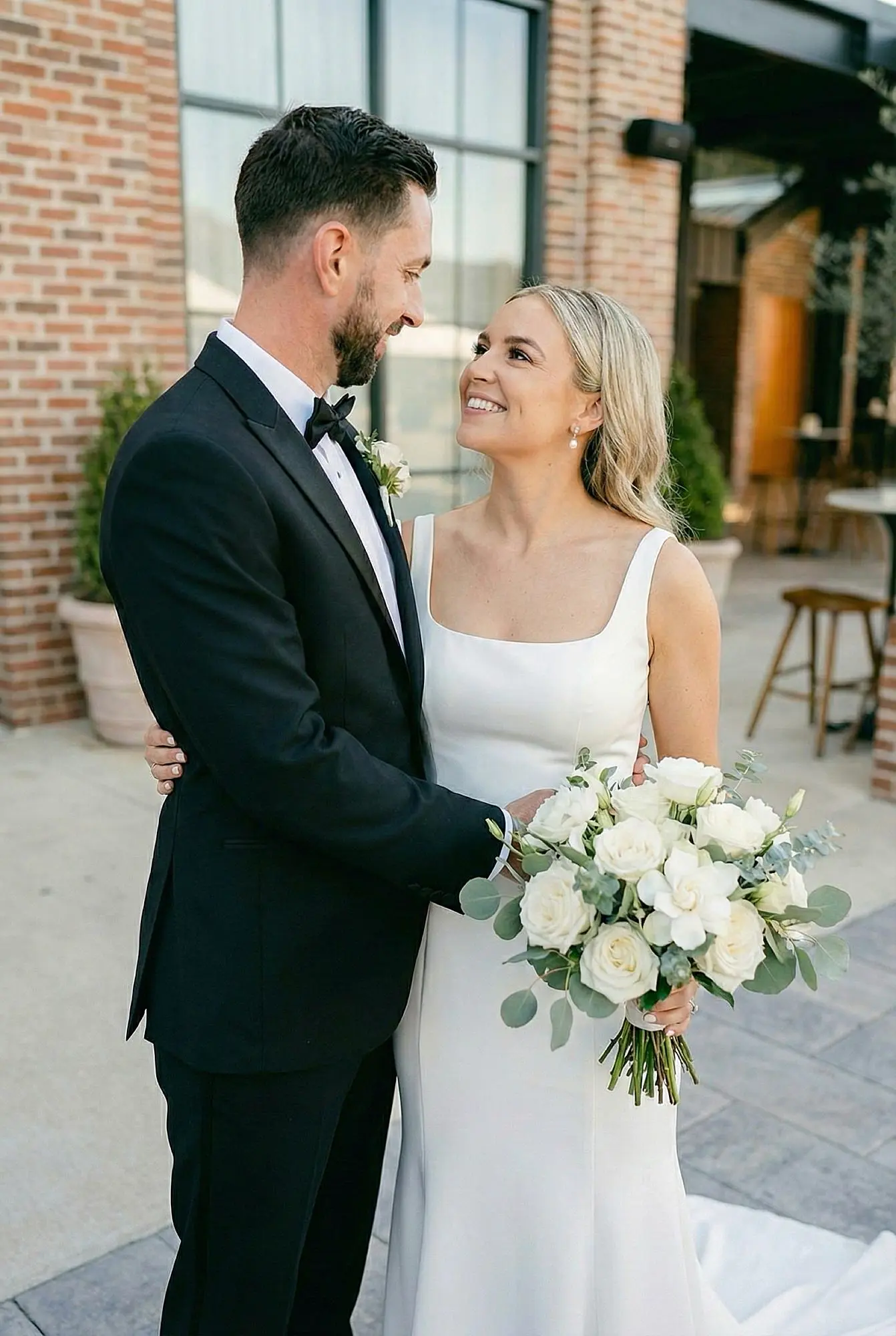 To help with the SEO for your Wedding Guide, here are the alt tags for the images in your gallery. I've focused on describing the elegant, clean aesthetic of the Valley Estate while keeping your brand keywords in mind.

Image Alt Tags
Alt Tag 1 (Top Left): Groom in a black tuxedo looking at the bride in a sleek white square-neck wedding dress holding a white peony bouquet at The Valley Estate.